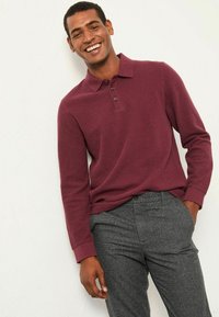 Bordeauxfarbenes Langarm-Polohemd mit drei Knöpfen am Kragen, kombiniert mit grauen gemusterten Hosen. Der Stoff wirkt strukturiert und lässig.