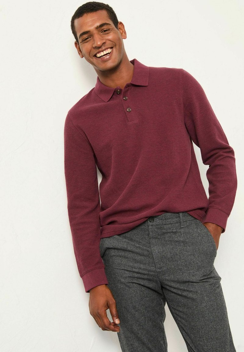 Bordeauxfarbenes Langarm-Polohemd mit drei Knöpfen am Kragen, kombiniert mit grauen gemusterten Hosen. Der Stoff wirkt strukturiert und lässig.
