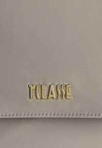 Logo in metallo dorato "T CLASSE" su tessuto beige con cuciture visibili lungo il bordo di un flap di una borsa o portafoglio.