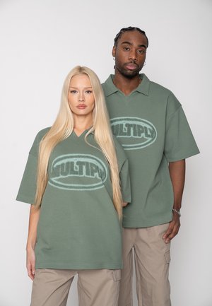 Doi adulți, o femeie și un bărbat, purtând tricouri verzi supradimensionate asortate cu logo-ul "MULTIPL" și pantaloni cargo bej, stând pe un fundal simplu.