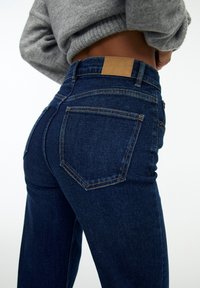 Jeans de mezclilla azul oscuro con una cintura alta, que presentan una etiqueta de cuero marrón en la parte posterior, una textura suave y un diseño clásico de cinco bolsillos.