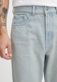 Mejores de mezclilla azul claro con un diseño clásico de cinco bolsillos, un ligero desvanecimiento y herrajes de metal con una superficie texturizada.