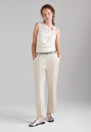 Giovane donna in piedi con le mani nelle tasche, indossa un top crema senza maniche, pantaloni beige con cintura a fantasia e sandali argento a ciabatta.