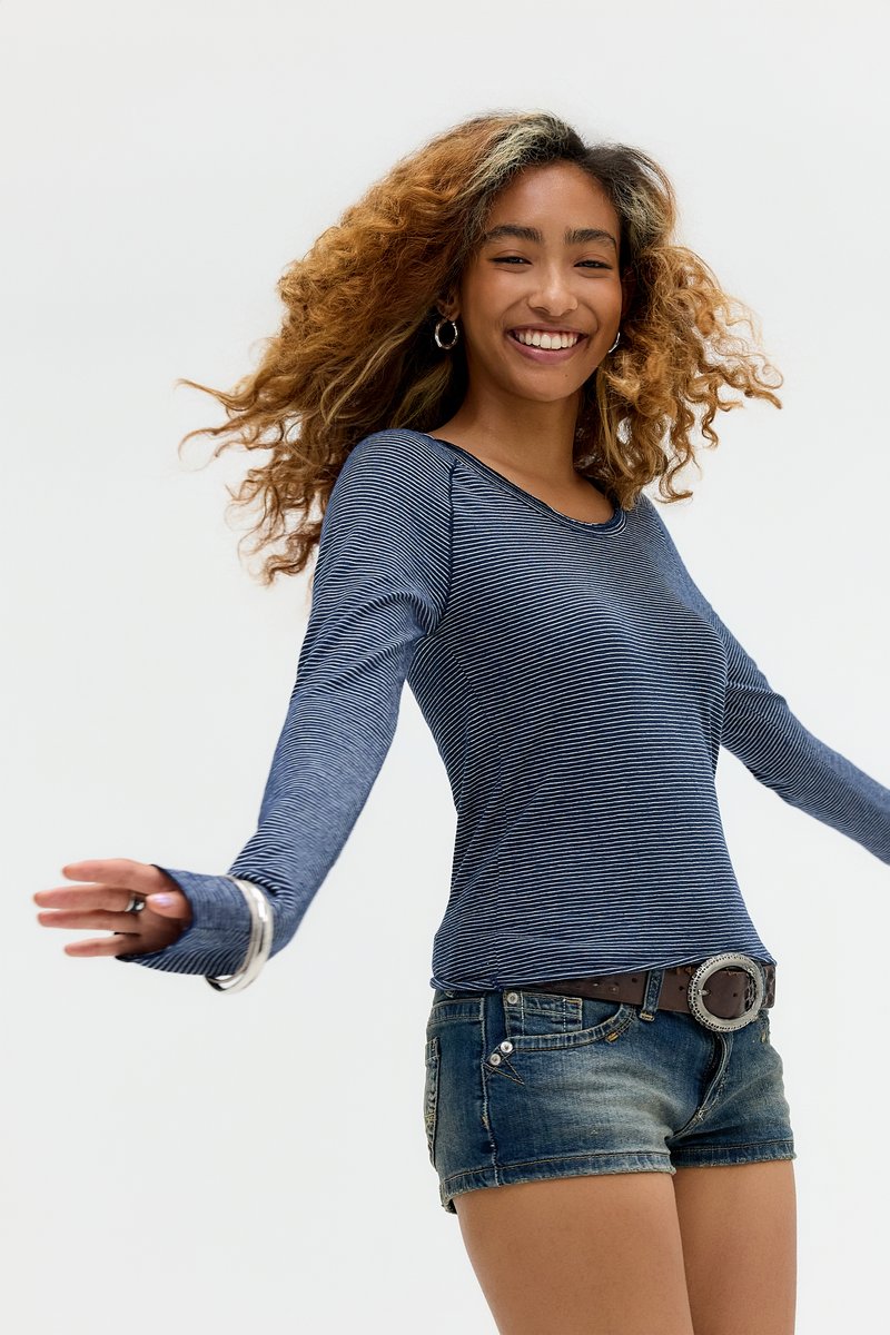 Ung kvinde med krøllet hår iført en langærmet, stribet top og denimshorts, smilende og snurrende mod en hvid baggrund.