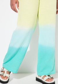 Lichte, getextureerde broek met een ombre-gradiënt van geel naar turquoise, gecombineerd met witte gespsandalen op een zwart plateau.