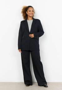 Schwarze karierte Blazer mit langen Ärmeln, kombiniert mit weit geschnittenen Hosen. Der Stoff hat ein strukturiertes Muster und wird von einem grauen Oberteil ergänzt.