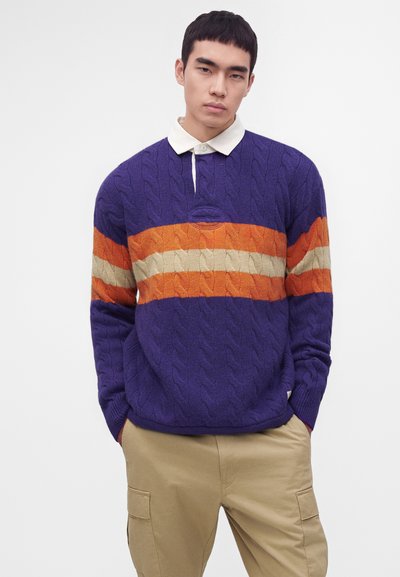 Jeune homme portant un pull torsadé violet avec des rayures orange et beige, une chemise blanche avec col, et un pantalon cargo beige, debout les mains dans les poches.