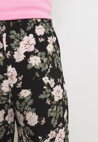 Pantalons noirs à motif floral avec de grandes fleurs roses et blanches accompagnées de feuilles vertes, taille élastique et tissu léger.