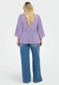 Lavendelkleurige gebreide top met een gestructureerd patroon en wijde mouwen, gecombineerd met lichtblauwe wijde jeans. Het achteraanzicht toont een open gebreid ontwerp.