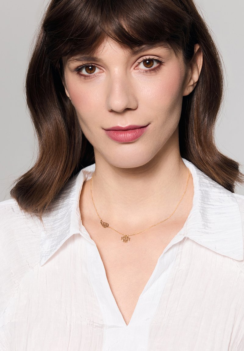 Złoty naszyjnik z dwoma wisiorkami: liściem i wzorem kwiatowym, noszony na białej koszuli z kołnierzykiem. Gładka tekstura z delikatnymi detalami.