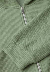 Felpa verde con zip e tessuto testurizzato, caratterizzata da una zip liscia in argento e polsini a coste. La superficie presenta un motivo a nido d'ape.