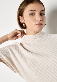 Strickoberteil in sanftem Creme mit hohem Hals, kurzen Ärmeln und einem strukturierten Rippmuster. Verfügt über einen subtilen Fall und eine lässige Passform.