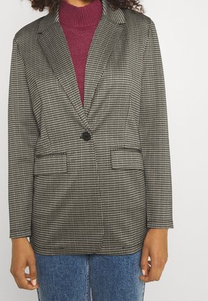 Blazer à carreaux noir et blanc avec un bouton unique, poches latérales et revers crantés, associé à un pull col roulé violet foncé et un jean bleu.