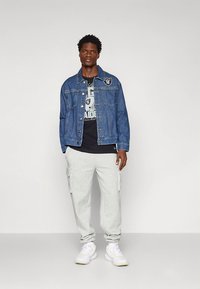 Τζιν μπουφάν με λογότυπο, φορεμένο πάνω από μαύρο T-shirt με σχέδιο, συνδυασμένο με γκρι παντελόνι cargo και λευκά αθλητικά παπούτσια.