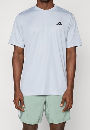 Mann trägt hellgraues, kurzärmeliges Sportshirt mit schwarzem Logo auf der Brust und hellgrünen Sportshorts.