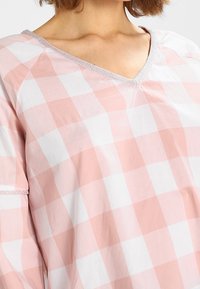 Blusa a cuadros en rosa claro y blanco con escote en V, con un adorno plateado en el cuello. Confeccionada en una tela suave y ligera.