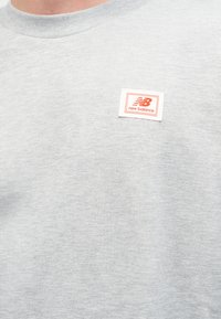 T-shirt gris à col rond avec un petit patch rectangulaire du logo New Balance en rouge sur le côté gauche de la poitrine.