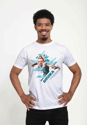 Hvid bomulds t-shirt med et grafisk motiv af en karakter i blå og beige nuancer, med teksten "AHSOKA" i fede, mørke bogstaver.