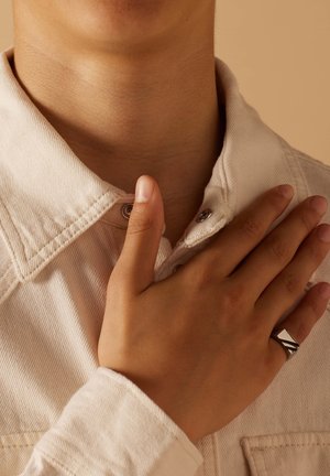 Silberring mit geometrischem Design auf einer Hand, die auf einem krämfarbenen Hemdkragen aus strukturiertem Stoff ruht. Hautfarbe ist sichtbar.