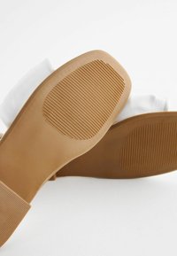 Due stivali in tessuto bianco con suole in gomma marrone chiaro che presentano pattern di grip a coste, mostrati dal fondo su uno sfondo bianco.