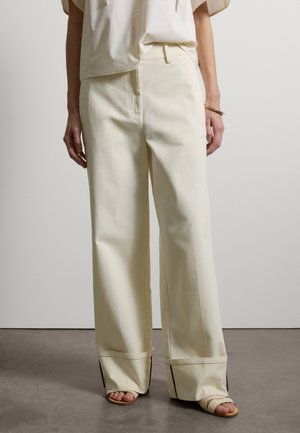 Person trägt hoch taillierte, weit geschnittene cremefarbene Hosen mit umgeschlagenen Säumen, kombiniert mit beigen Sandalen mit offenen Zehen und einem weißen, in die Hose gesteckten Oberteil.