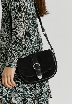 Kobieta ubrana w wzorzystą sukienkę z długim rękawem trzyma czarną skórzaną torebkę na ramię z srebrną klamrą i regulowanym paskiem.