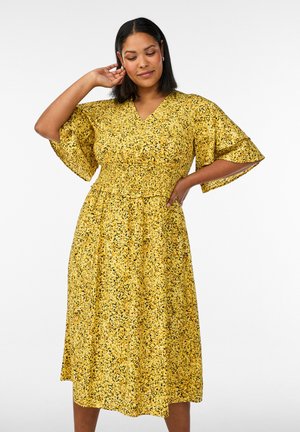 Woman posing with hand touching hair and other on hip, wearing a yellow floral midi dress with flutter sleeves.