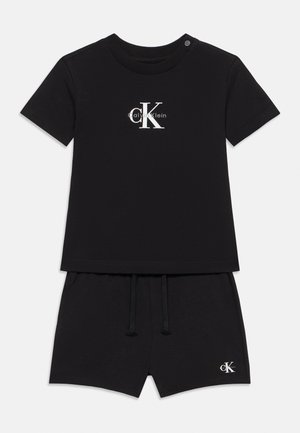 Schwarzes T-Shirt- und Shorts-Set in Kleinkindgröße mit weißem Calvin Klein "cK" Logo auf der Brust und am Saum der Shorts, mit Knopf an der Schulter des Shirts.