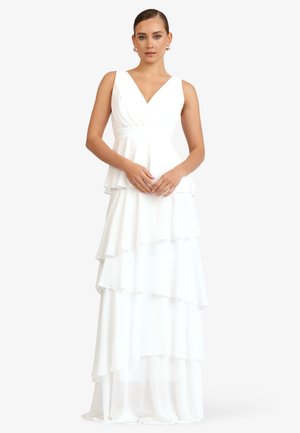 Kobieta ubrana w białą, warstwową sukienkę bez rękawów z dekoltem w kształcie litery V, stojąca ze splecionymi rękami na tle jednolitego tła.