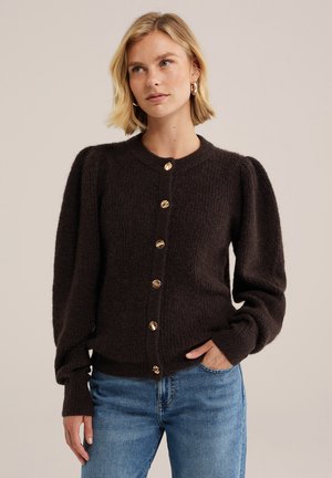 Frau mit kurzen blonden Haaren, die einen dunkelbraunen, geknöpften Pullover mit Puffärmeln und blaue Jeans trägt, steht mit einer Hand in der Tasche.