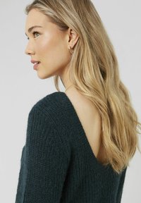 Pull en maille vert foncé, présentant un dos en V profond et un tissu texturé. La vue de côté met en avant de longs cheveux et des bijoux d'oreilles en or minimalistes.