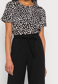 Mujer con una blusa de manga corta con un patrón en blanco y negro metida dentro de pantalones negros de tiro alto, con un cinturón de tela atado.