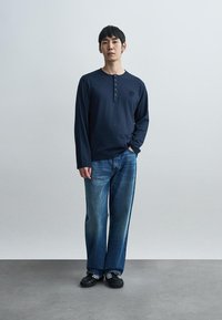 Μπλε ναυτικό μακρυμάνικο πουκάμισο henley με κουμπιά, σε συνδυασμό με ξεβαμμένο μπλε τζιν. Το μοντέλο στέκεται σε γκρι πάτωμα μπροστά από μιας ουδέτερης απόχρωσης φόντο.