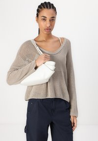 Beiger Strickpullover mit lockerer Passform, kombiniert mit marineblauen Cargo-Hosen. Weße Lederhandtasche, die in einer Hand gehalten wird, mit einem einzigartigen gefalteten Design.