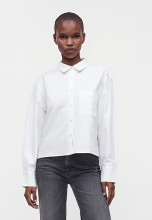 Persona con el pelo corto que lleva una camisa blanca de manga larga con botones y vaqueros gris oscuro, de pie frente a un fondo liso.