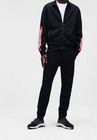 Chándal negro con chaqueta de cremallera y pantalones a juego. La chaqueta presenta texto gráfico rojo en las mangas, combinado con zapatillas deportivas negras.