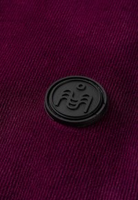 Czarne gumowe logo na fakturowanej, głęboko purpurowej tkaninie z poziomymi rowkami. Logo przedstawia abstrakcyjne kształty w granicach okrągłej ramki.