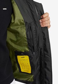 Musta ulkotakki, jossa sileä vihreä vuori. Takissa on vetoketjullinen tasku ja keltataustainen lappu, jossa on symboleja.