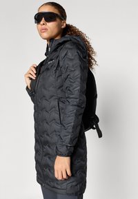 Čierna prešívaná dlhá bunda so zipsom a kapucňou. Štruktúrovaná tkanina. Nosená s čiernym batohom a veľkými slnečnými okuliarmi.