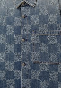 Vêtement en denim avec motif damier texturé, boutons-pression métalliques, col pointu et poche poitrine rectangulaire avec coutures visibles.