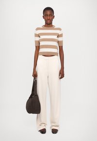 Braun-weiß gestreifter, cropped Pullover, cremefarbene weite Hose, dunkelbraune Tasche und Schuhe. Modell steht mit den Händen an den Seiten nach vorne gerichtet.