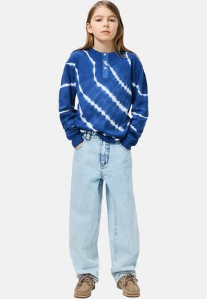 Camisa de manga larga con teñido anudado azul y patrones blancos, abertura con botones, combinada con vaqueros anchos azul claro y zapatos marrones.