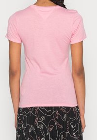 Žena v jednoduchém světle růžovém tričku s krátkým rukávem a černé sukni s abstraktními bílými a růžovými liniemi tváře, zobrazená zezadu.