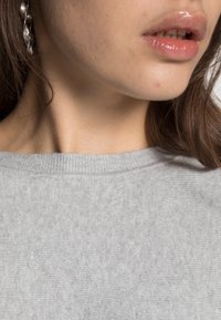 Pull gris avec un col côtelé, texture douce et coupe décontractée, associé à des boucles d'oreilles discrètement brillantes visibles près du cou.