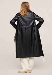 Sort læderlang frakke med lige snit, rygbånd og lange ærmer. Sattes sammen med cremede bukser og let fodtøj.