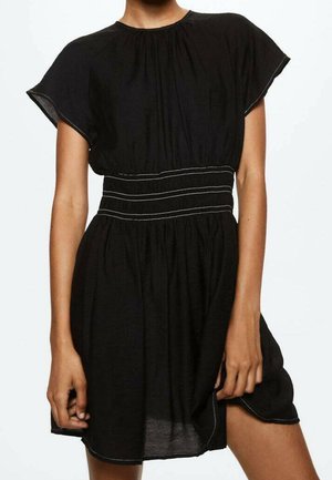Schwarzes Kleid mit kurzen Ärmeln, geraffter Taille und weißen Ziernähten, getragen von einer stehenden Person, Hände ruhen nahe am Saum.