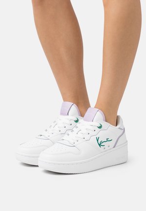 Baskets basses blanches avec signature verte sur le côté, accents lavande sur la languette et le talon, portées sur des jambes nues sur un fond blanc.