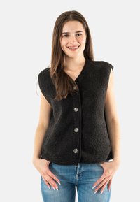 Chaleco de forro polar negro con una textura suave, diseño sin mangas, escote en forma de v y cinco botones plateados en el frente. Llevado sobre jeans azules.