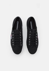 Svarta canvas-sneakers med vita gummisulor, med silverfärgade ögla, rund tå och strukturerad yta. Varumärkeslogotyp synlig på insoles.