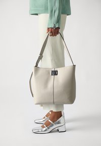 Duża beżowa skórzana torba typu tote o zarysowanej konstrukcji, z zapięciem z srebrnymi elementami i długim regulowanym paskiem. Widoczne srebrne buty na obcasie.